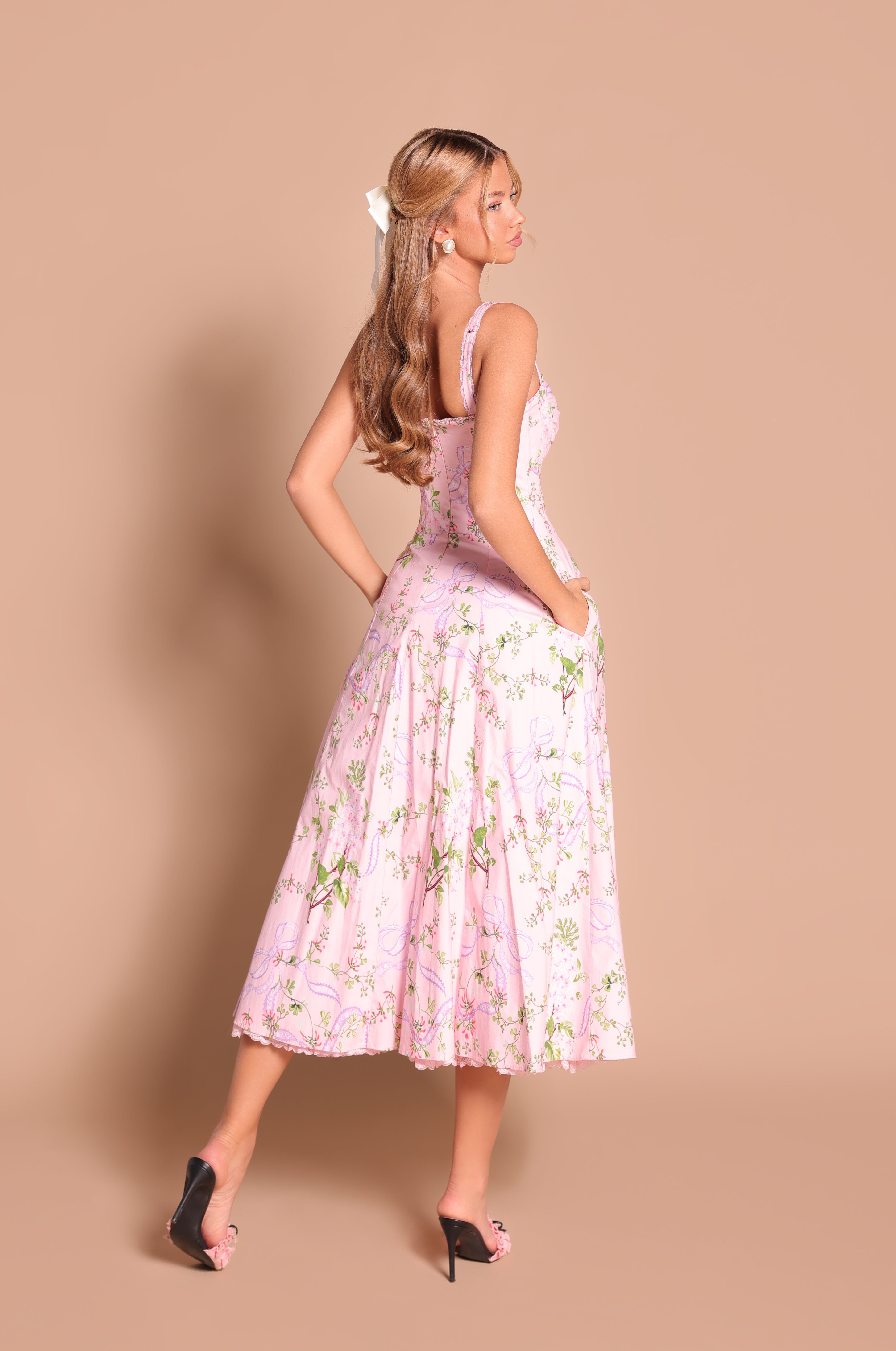 A woman stands against a plain beige background wearing the Celenia Blush Pink Floral Casual Midi Dress with pockets, paired with black heeled sandals and styled with long, wavy hair.